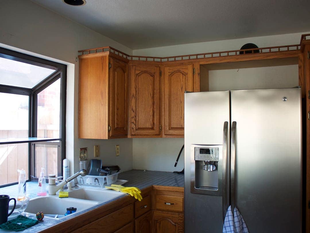 Existing 1990s kitchen: oak cabinets, tile countertop, double sink under a corner garden window, side-by-side refrigerator